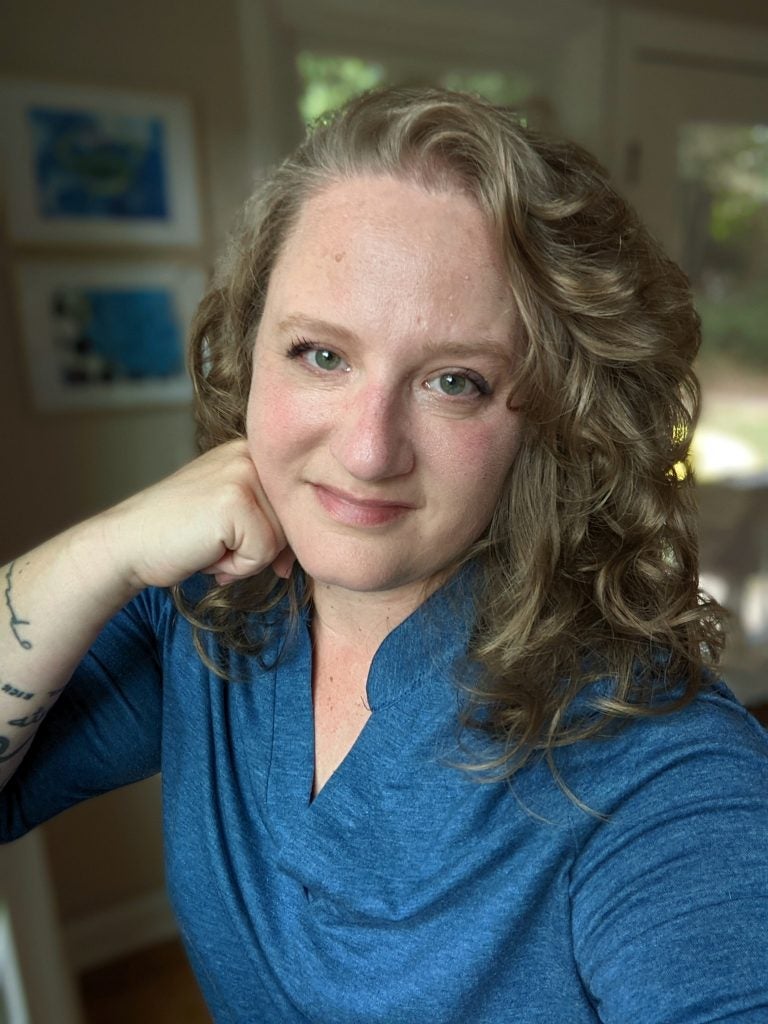 Devra Thomas, Digital Editor. A light skinned white woman with dark blonde wavy hair, wearing a blue shirt. 