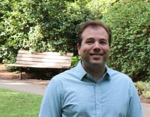 James Tate Hill, a white man with short brown hair, smiles, wearing a light blue checked button down shirt. standing inf front of green hedges and a brown bench.