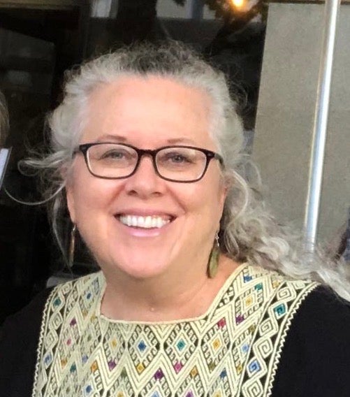 A gray-haired woman wearing glasses and a black dress with intricate colorful embroidery around the neckline.