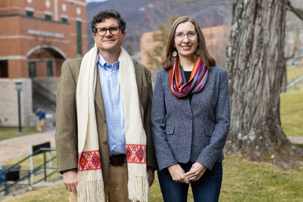 A white man with black hair and black glasses, wearing a brown sportcoat and blue buttonup shirt, standing beside a white woman with brown hair and glasses, wearing a blue tweed jacket and a colorful scarf. They are standing outside in front of a tree and a brick building.