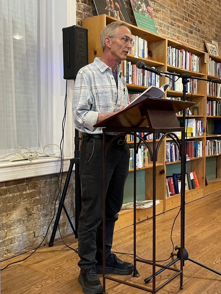 Jeffrey, a white man with grey hair and glasses, wearing a white and grey plaid shirt and black pants, standing in front of a lectern and microphone, in a bookstore.