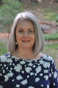 Ashley Memory, grey bob hair, blue top with white daisies on it. She is standing outside. 