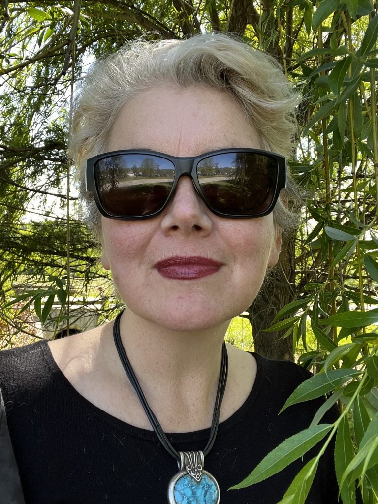 Laura Hope-Gill, wearing black sunglasses and a black shirt, with a blue necklace and short grey hair. She is outdoors in front of green tree leaves.
