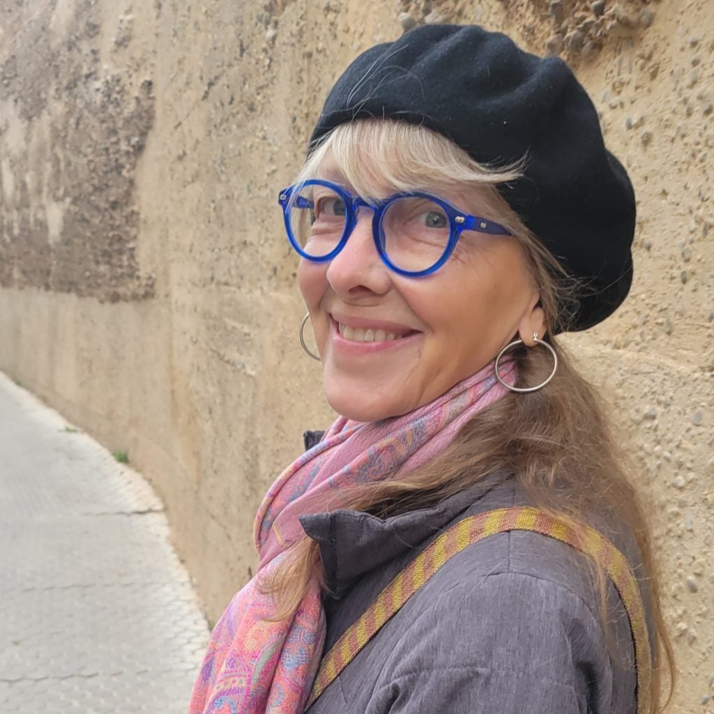 Dawn Reno Langley, wearing bright blue eyeglasses and a black beret, with a pink scarf. 