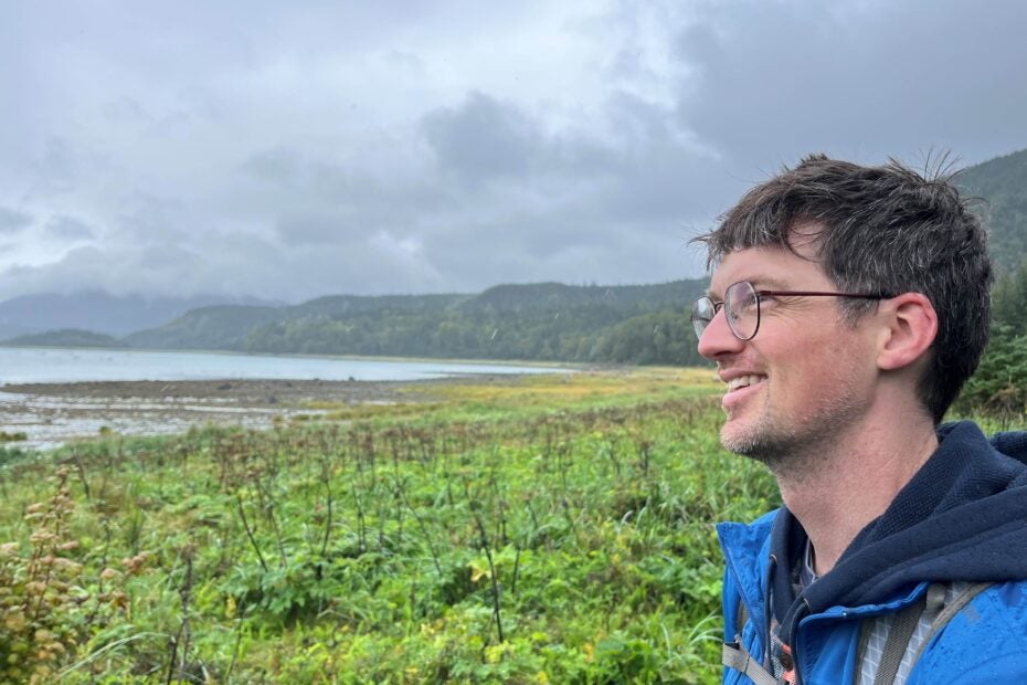 Evan, a white man with brown hair and eyeglasses, wearing a blue jacket with a black hoodie, standing in front of green marsh and grey cloudy sky