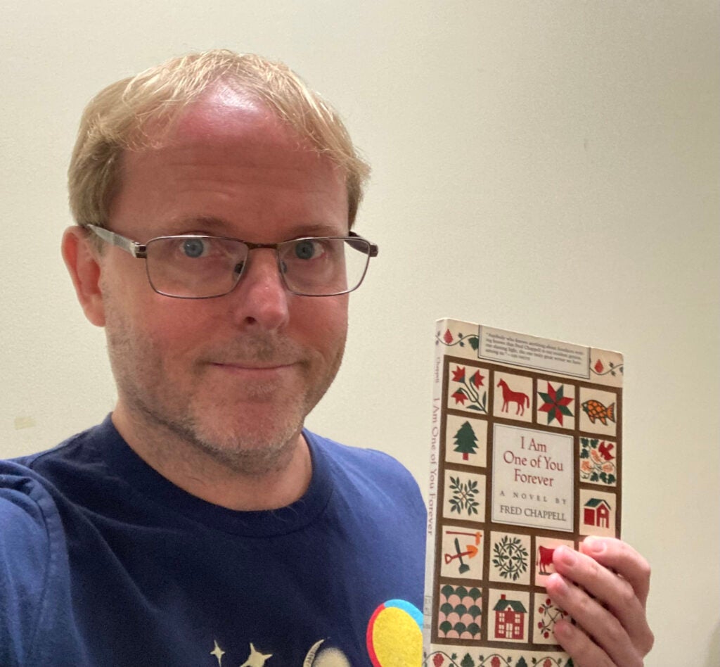 Randall, a white man with blond hair and glasses, wearing a blue shirt, holds a copy of Fred Chappell's novel I Am One of You Forever.