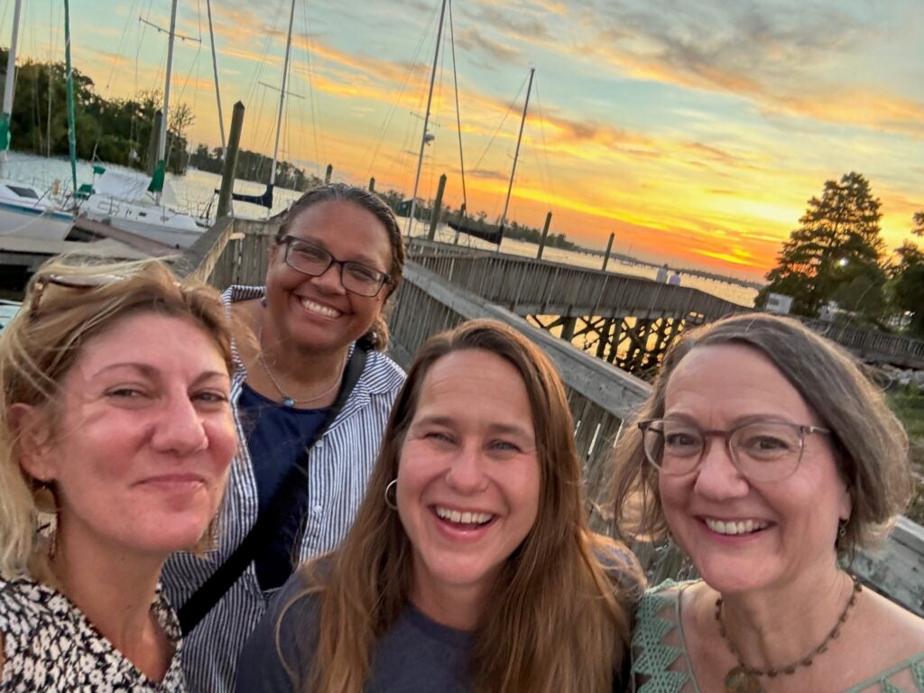 four women, standing on a dock, in front of an orange sunset, with sailboats in the background